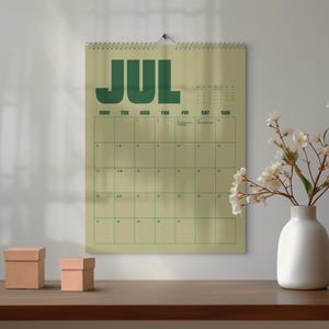 May include: A wall calendar for July in green and beige. The word "JUL" is printed in large, bold, green letters at the top. The calendar grid is also green. Two small, brown cardboard boxes sit on a wooden surface.