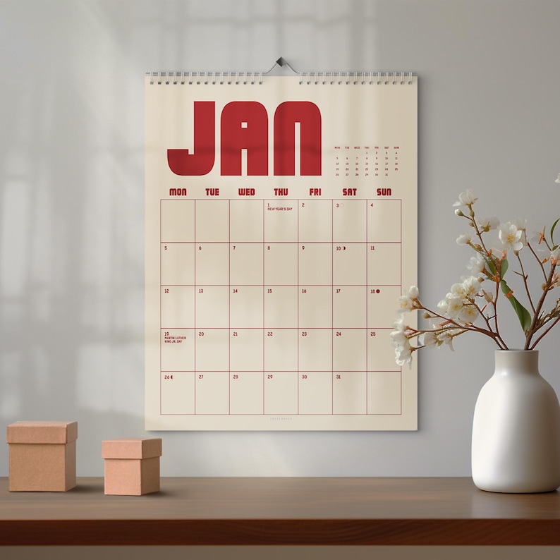May include: A wall calendar for January with a cream background and red lettering. The word "JAN" is at the top, with days of the week below. The calendar is hanging on a wall, with a vase of flowers and two small boxes on a wooden surface.
