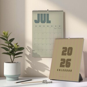 May include: Two calendars, one hanging and one standing, are displayed. The hanging calendar shows "JUL" in large blue letters. The standing calendar reads "2026 CALENDAR" in brown. A potted plant and pencils are in the foreground.