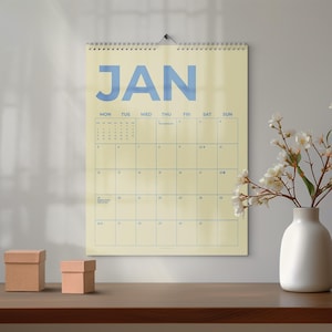 May include: A cream-coloured wall calendar for January, with blue text. The word "JAN" is at the top, with the days of the week below. A white vase with white flowers and two small brown boxes are on a wooden surface.