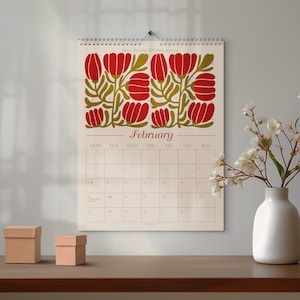 Könnte beinhalten: Ein Wandkalender für Februar mit einem floralen Design in Rot und Olivgrün. Der Kalender hängt an einer Wand über einem Holztisch. Zwei kleine, quadratische Pappschachteln stehen auf dem Tisch, und rechts steht eine weiße Vase mit Blumen.