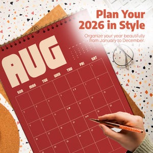 May include: A red 2026 calendar with the month of August displayed. The calendar is open, showing the days of the month. The text "Plan Your 2026 in Style" is at the top. A hand is holding a pen, ready to write on the calendar.