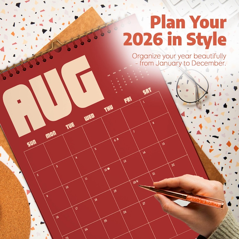 May include: A red 2026 calendar with the month of August displayed. The calendar is open, showing the days of the month. The text "Plan Your 2026 in Style" is at the top. A hand is holding a pen, ready to write on the calendar.