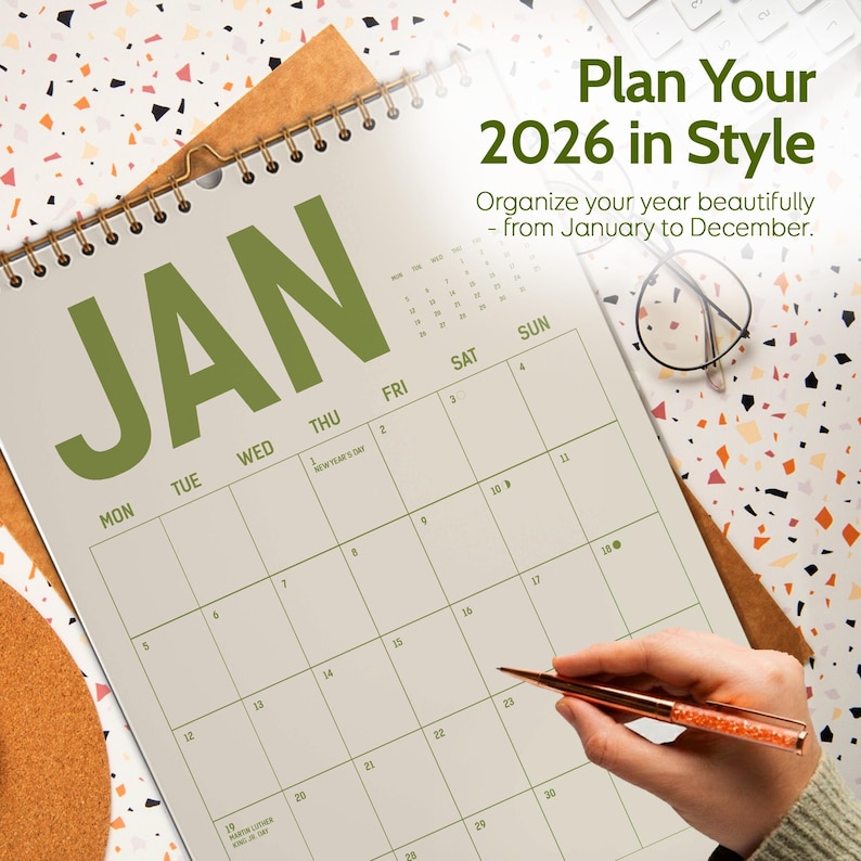 May include: A large, open calendar for the year 2026, with the month of January prominently displayed. The calendar is open to the first week of the month. The text "Plan Your 2026 in Style" is at the top. A hand is holding a pen.