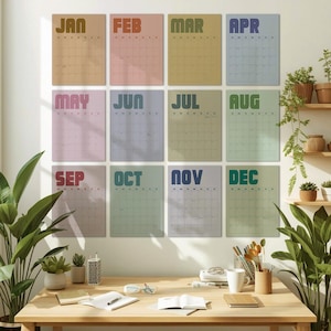 May include: A set of twelve monthly calendars, each with a different color background and month name. The months are displayed in a retro font. The calendars are hanging on a white wall above a wooden desk with plants.