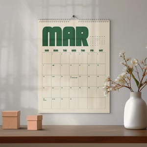 Puede incluir: Un calendario de pared para el mes de marzo con grandes letras de bloque de color verde oscuro en la parte superior. La cuadrícula del calendario está impresa en verde oscuro sobre un fondo crema, mostrando los días y fechas, incluido San Patricio.