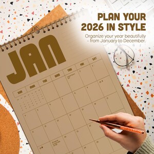 May include: A spiral-bound 2026 calendar open to January. The calendar is tan with dark brown text and dates. The text "PLAN YOUR 2026 IN STYLE" is at the top. A hand holds a pen, and glasses rest on the patterned surface.