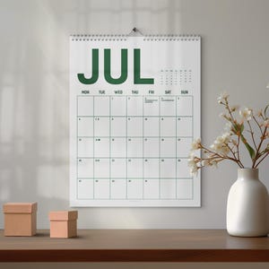 Könnte beinhalten: Ein weißer Wandkalender für Juli mit grünen Buchstaben und Rasterlinien, der an einer Wand hängt. Eine weiße Vase mit weißen Blumen und zwei kleine braune Schachteln stehen auf einer Holzoberfläche unter dem Kalender.