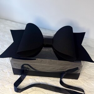 Pode incluir: Uma caixa de presente preta com um grande laço decorativo no topo. A caixa tem uma janela de plástico transparente e alças de fita azul escura. O laço é feito de papel ou cartão preto espesso. A caixa está sobre uma superfície branca texturizada.