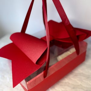 Könnte beinhalten: Eine rote Geschenkbox mit einem transparenten Fenster, einer großen Schleife und einem Griff aus rotem Band. Die Box steht auf einer weißen Oberfläche. Die Box ist wahrscheinlich für ein Geschenk.