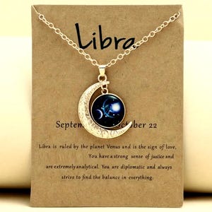 Può includere: Una collana color argento con un ciondolo a forma di mezzaluna e un centro rotondo blu scuro con un design celeste. La collana è appesa a una carta con la parola "Libra" e il testo sul segno zodiacale.