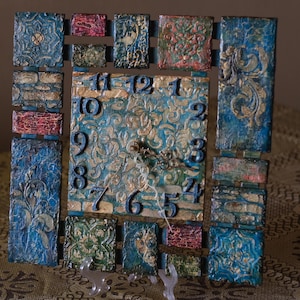 Op de afbeelding: Vierkante wandklok met een textuur, sierlijk ontwerp. De wijzerplaat is lichtblauw met gouden accenten en zwarte cijfers. Het frame bestaat uit verschillende rechthoekige stukken in tinten blauw, rood en groen.