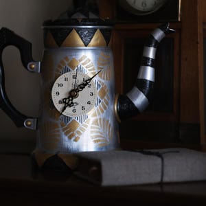 Op de afbeelding: Een decoratieve theepotvormige klok met een zilverkleurige behuizing, zwarte handgreep en tuit. De wijzerplaat is wit met zwarte cijfers en wijzers. Gouden en zwarte geometrische patronen sieren de theepot. Een klein, rechthoekig, grijs object ervoor.