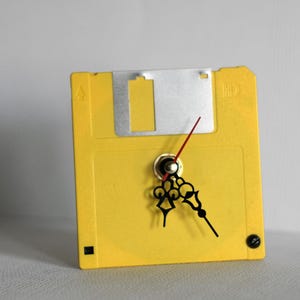 Op de afbeelding: Een gele floppy disk hergebruikt als klok. De klok heeft zwarte wijzers, een rode secondewijzer en een zilveren metalen plaat. De klok is een uniek decoratief item.