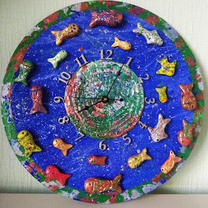 Op de afbeelding: Een ronde, handgeschilderde wandklok met een blauwe achtergrond en kleurrijke visaccenten. De wijzerplaat heeft een getextureerd, groen en rood midden, zilveren cijfers en zwarte wijzers. De klok is versierd met diverse visfiguren.