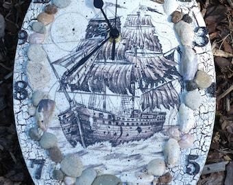 Puede incluir: Reloj de pared ovalado con temática náutica. La esfera del reloj presenta una ilustración en tonos sepia de un velero. El borde del reloj está decorado con pequeñas piedras y conchas marinas. Las manecillas del reloj son negras.