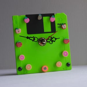May include: A unique, handmade clock crafted from a bright green floppy disk. The clock features black clock hands, a gold-colored center, and colorful fruit-shaped embellishments. The clock face has the text "FUJIFILM DOS".