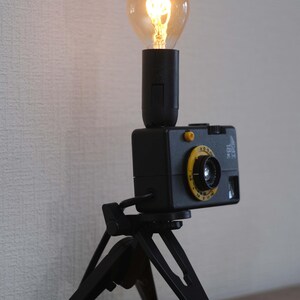Könnte beinhalten: Eine Tischlampe bestehend aus einem schwarzen Vintage-Kameragehäuse, das auf einem kleinen schwarzen Stativ montiert ist. Eine schwarze zylindrische Fassung ragt aus der Oberseite der Kamera und hält eine leuchtende, runde Glühbirne.