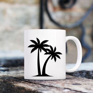 May include: White ceramic mug featuring a black silhouette of two palm trees. The mug has a curved handle and is set against a blurred background of wood and a wall.