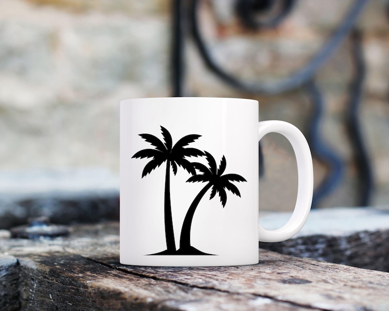 May include: White ceramic mug featuring a black silhouette of two palm trees. The mug has a curved handle and is set against a blurred background of wood and a wall.