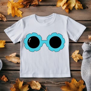 May include: White t-shirt featuring a cartoon design of sunglasses with black lenses and a light blue floral frame. The shirt is displayed on a wooden surface with autumn leaves and a pair of white sneakers.