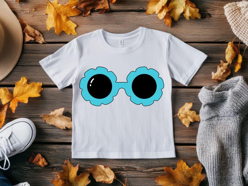 May include: White t-shirt featuring a cartoon design of sunglasses with black lenses and a light blue floral frame. The shirt is displayed on a wooden surface with autumn leaves and a pair of white sneakers.
