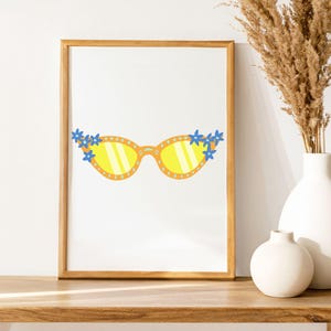 May include: A framed print featuring a whimsical illustration of yellow sunglasses adorned with blue flowers. The sunglasses have yellow lenses and are set against a white background. The frame is a warm, light brown color. The print is displayed on a wooden surface.