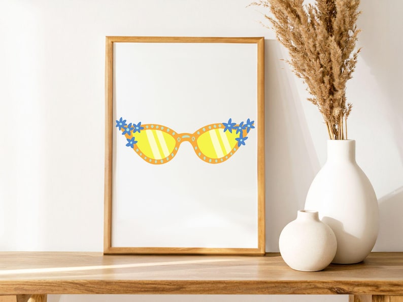 May include: A framed print featuring a whimsical illustration of yellow sunglasses adorned with blue flowers. The sunglasses have yellow lenses and are set against a white background. The frame is a warm, light brown color. The print is displayed on a wooden surface.