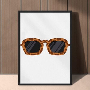 May include: A framed print featuring a pair of sunglasses with a brown and black leopard print pattern. The sunglasses have dark lenses and a rectangular shape. The artwork is displayed against a white background.