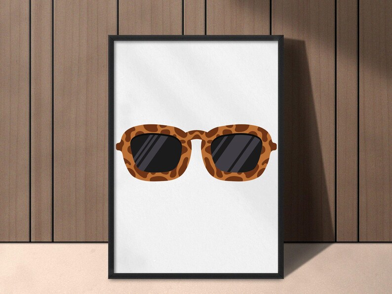 May include: A framed print featuring a pair of sunglasses with a brown and black leopard print pattern. The sunglasses have dark lenses and a rectangular shape. The artwork is displayed against a white background.