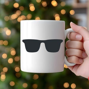 May include: White ceramic mug featuring a black sunglasses graphic. The mug has a curved handle and is held by a person. The background is blurred with green and gold bokeh lights, suggesting a festive or holiday setting.
