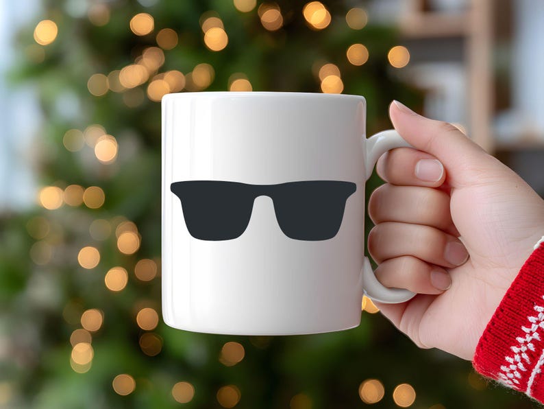 May include: White ceramic mug featuring a black sunglasses graphic. The mug has a curved handle and is held by a person. The background is blurred with green and gold bokeh lights, suggesting a festive or holiday setting.