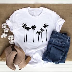 May include: White t-shirt with a black palm tree graphic. The shirt is paired with blue denim jeans and tan ankle boots. The items are arranged on a neutral-toned surface, creating a casual, everyday look.