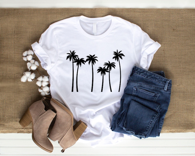 May include: White t-shirt with a black palm tree graphic. The shirt is paired with blue denim jeans and tan ankle boots. The items are arranged on a neutral-toned surface, creating a casual, everyday look.