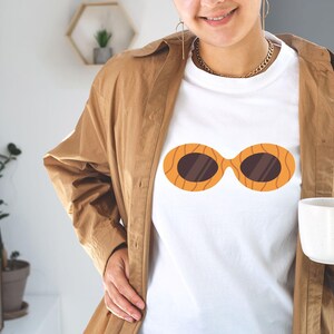 May include: White t-shirt featuring a graphic of orange sunglasses with brown lenses. The sunglasses design has a wood grain pattern. The person is wearing a brown jacket and holding a white mug.