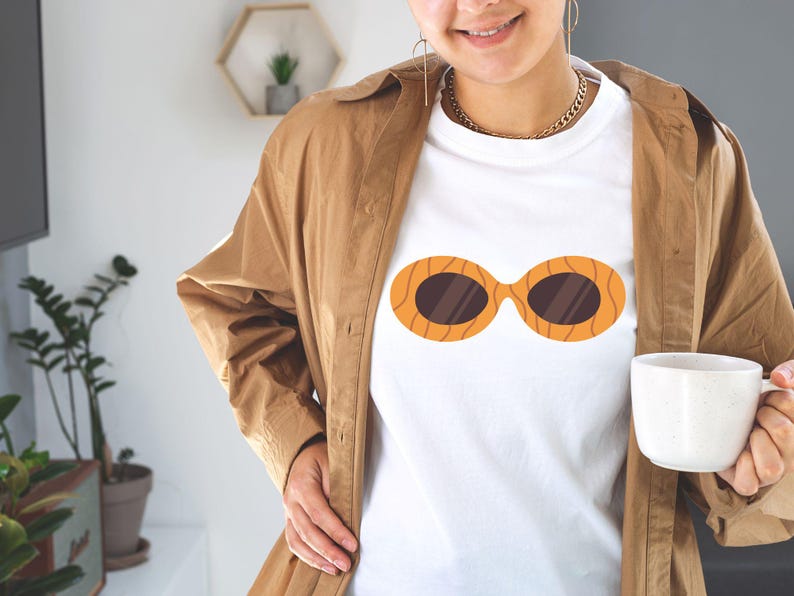 May include: White t-shirt featuring a graphic of orange sunglasses with brown lenses. The sunglasses design has a wood grain pattern. The person is wearing a brown jacket and holding a white mug.