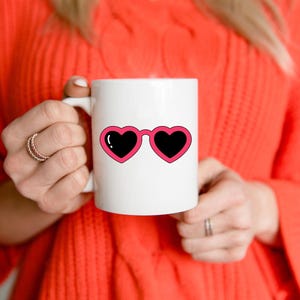 May include: White ceramic mug featuring a pink heart-shaped sunglasses design with black lenses. The mug is held by a person wearing an orange sweater. The design is a fun and playful accessory.