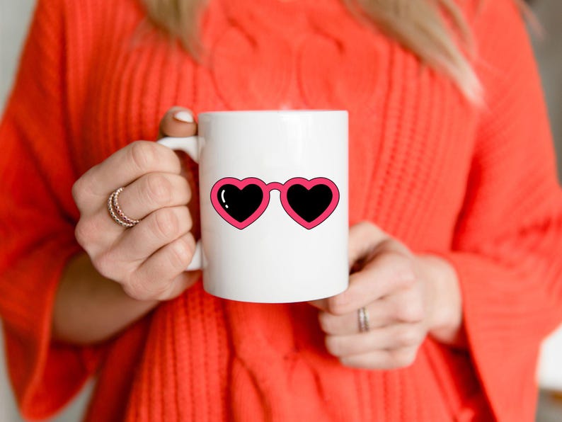 May include: White ceramic mug featuring a pink heart-shaped sunglasses design with black lenses. The mug is held by a person wearing an orange sweater. The design is a fun and playful accessory.