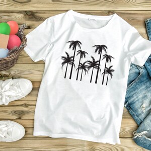 May include: White t-shirt with a black palm tree graphic. The shirt is displayed on a wooden surface with a pair of white sneakers, a basket of colorful Easter eggs, and a pair of blue jeans.