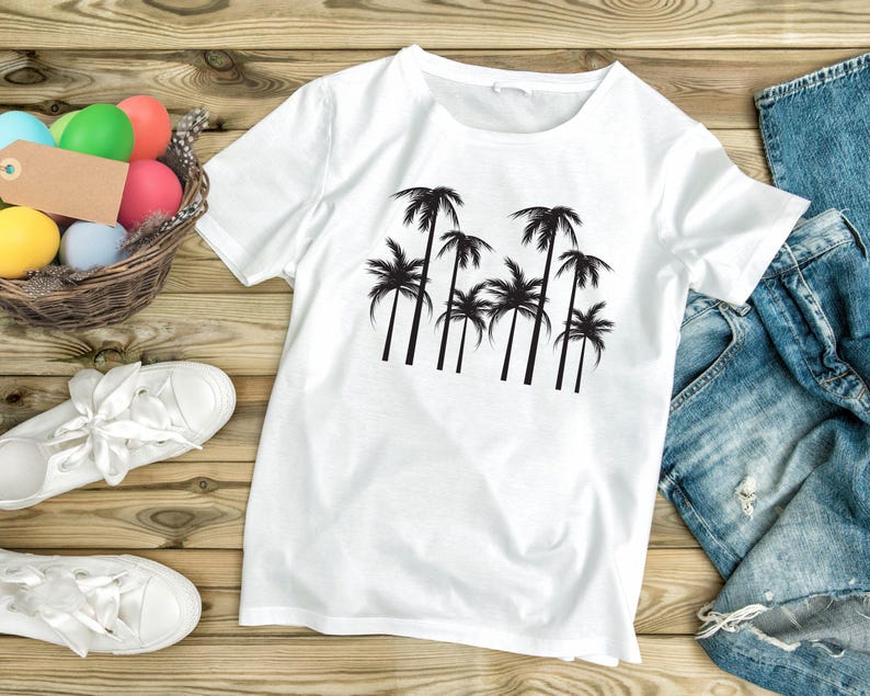 May include: White t-shirt with a black palm tree graphic. The shirt is displayed on a wooden surface with a pair of white sneakers, a basket of colorful Easter eggs, and a pair of blue jeans.