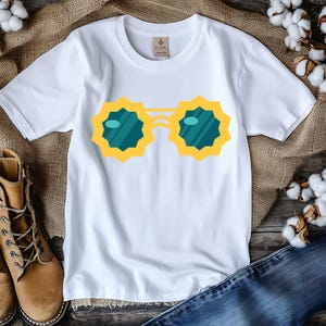 May include: White t-shirt featuring a yellow and teal sunglasses graphic. The sunglasses have a unique, starburst-like design. The shirt is displayed with brown boots and blue jeans, suggesting a casual, everyday style.