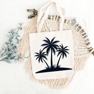 May include: Cream-colored tote bag featuring a black silhouette of three palm trees on a small island. The bag has two handles and is displayed on a white wooden surface with decorative elements.