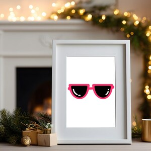 May include: A framed print featuring a pair of sunglasses with a bright pink frame and black lenses. The print is displayed on a table with holiday decorations, including a gold mug and a Christmas garland.