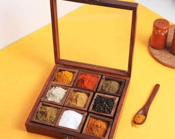 May include: A wooden spice box with a glass lid, filled with various colorful spices. A small wooden spoon holds a portion of yellow spice. The box is on a yellow surface, with a pair of salt and pepper shakers in the background.