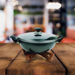 Op de afbeelding: Een donkergrijze kookpot met een glazen deksel en zwarte handvatten, rustend op een houten onderzetter. De pot is ontworpen voor gebruik op het fornuis. De onderzetter is gemaakt van hout en heeft een uniek, decoratief ontwerp.