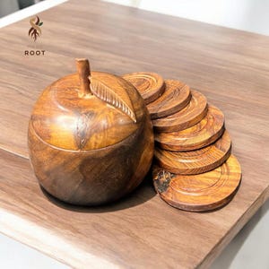 May include: A wooden apple-shaped container with a leaf detail on top, accompanied by a stack of matching wooden coasters. The container and coasters are made of a warm-toned wood, and the text "MADE ROOT" is visible.