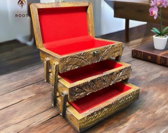 May include: A wooden jewelry box with three pull-out drawers and a hinged top. The interior is lined with red fabric. The box has intricate carved floral designs and is displayed on a wooden surface. The box is made by Made Root.