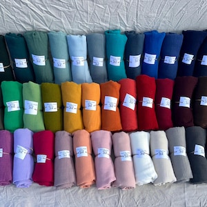 May include: A selection of rolled scarves in a spectrum of colours, such as green, blue, yellow, orange, red, and purple. Each scarf is secured with a white label and a brown string. The scarves are displayed on a white surface.