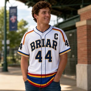 Pode incluir: Camisola de beisebol branca com a palavra "BRIAR" e o número "44" em azul marinho. A camisola tem detalhes em vermelho, amarelo e azul. O número "4" está em ambas as mangas. A camisola é usada por uma pessoa.
