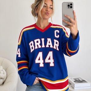 Könnte beinhalten: Blaues Langarm-Trikot mit dem Wort "BRIAR" und der Nummer "44" in Weiß. Das Trikot hat rote, gelbe und weiße Streifen an den Ärmeln und am Saum. Es hat einen roten V-Ausschnitt und den Buchstaben "C" oben rechts.